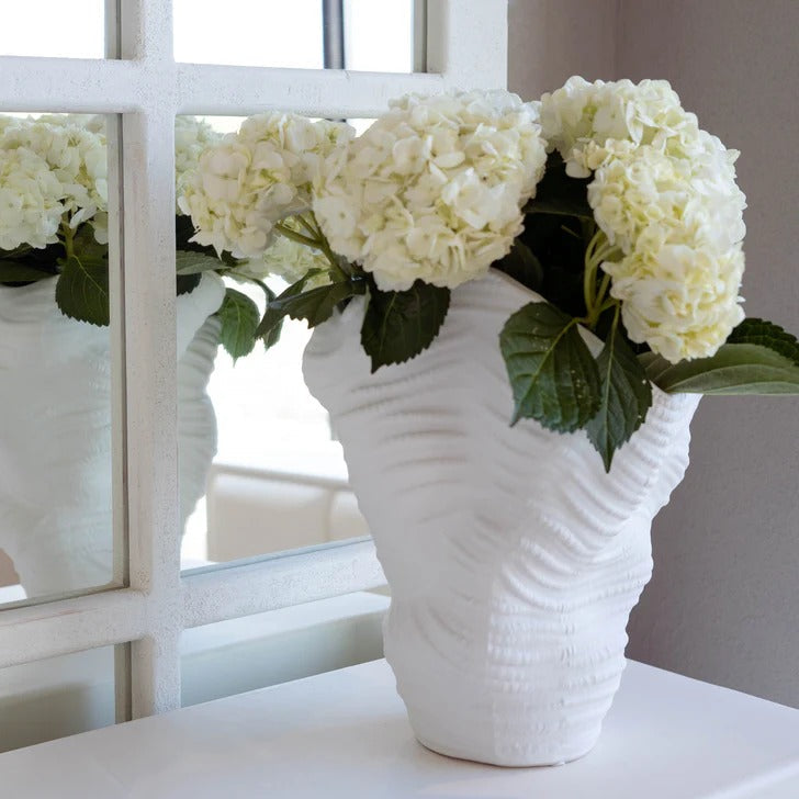White textured vase with white flowers on a table in a room with a mirror.