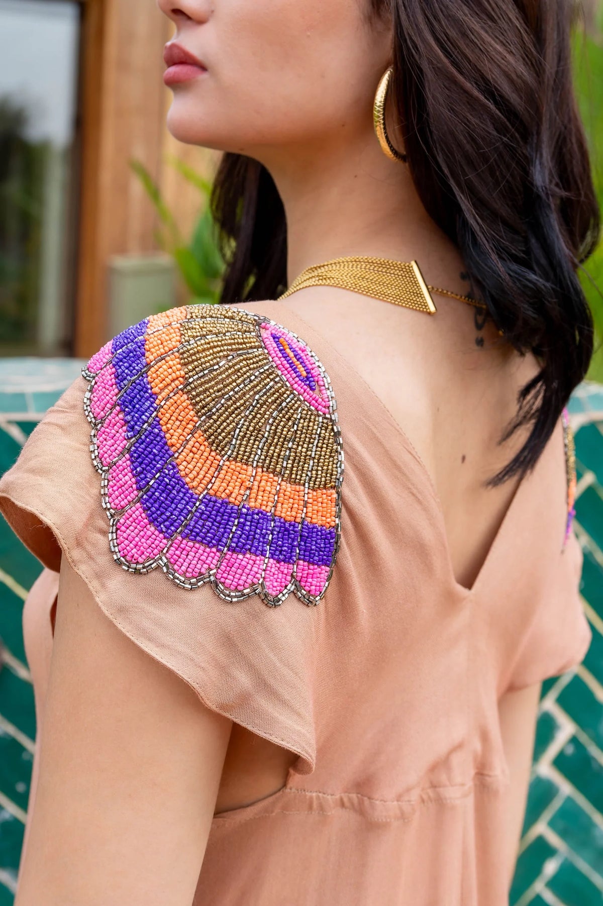 Woman wearing a peach-colored top with a colorful beaded back design, gold necklace, and green background.