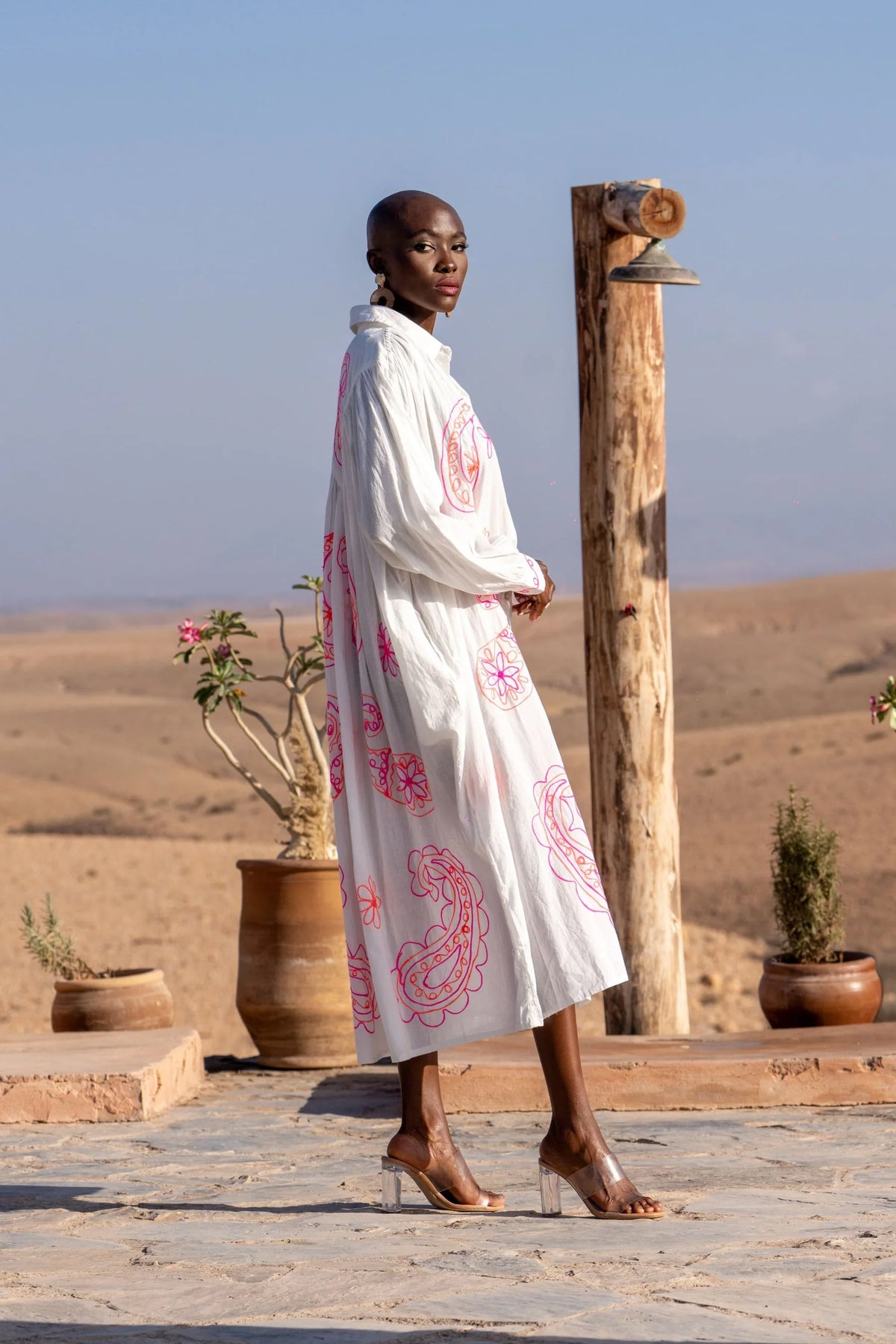 Woman in a white dress with pink patterns standing in a desert-like setting with wooden posts and plants.