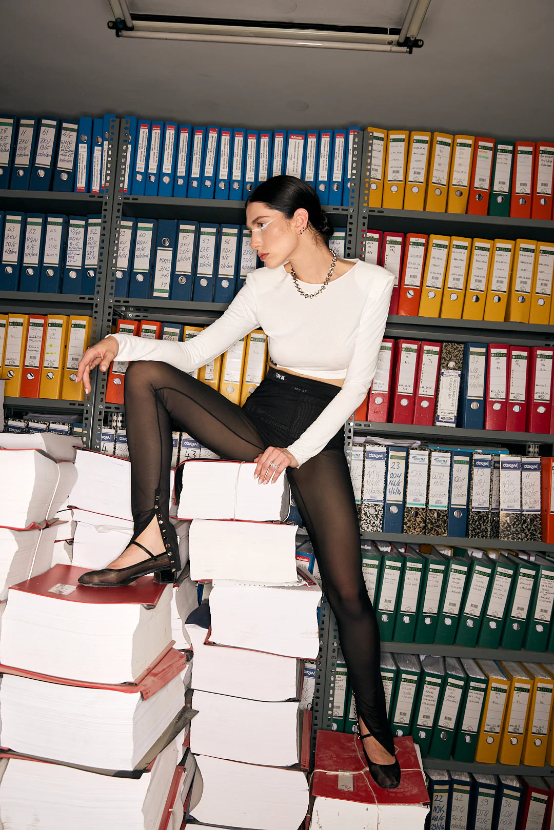 Woman sitting on a stack of books in an office setting with shelves of binders behind her