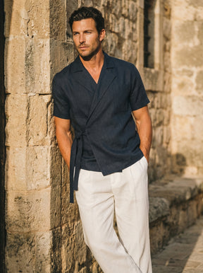 Man wearing a navy shirt and white pants standing against a stone wall.