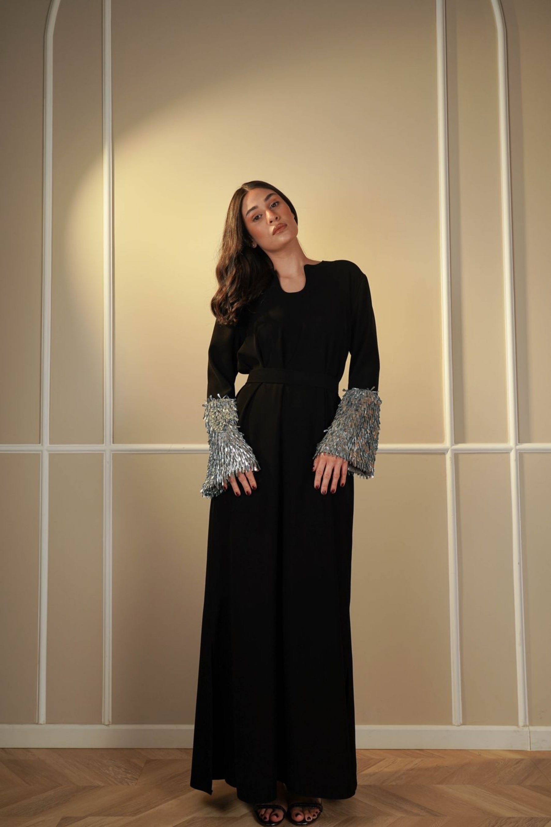 Woman wearing a long black dress with silver cuffs against a beige wall.