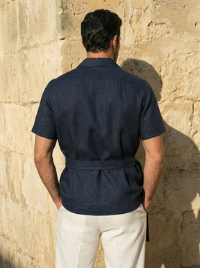 Man wearing a navy shirt and white pants against a stone wall.