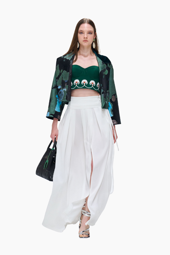 Model wearing a green top with floral design, white pants, and a black jacket on a white background