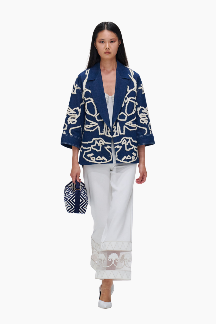 Woman wearing a navy blue jacket with white patterns and white pants on a white background