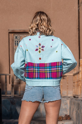 Person wearing a light blue denim jacket with colorful plaid pattern and floral embroidery, standing against a beige wall.
