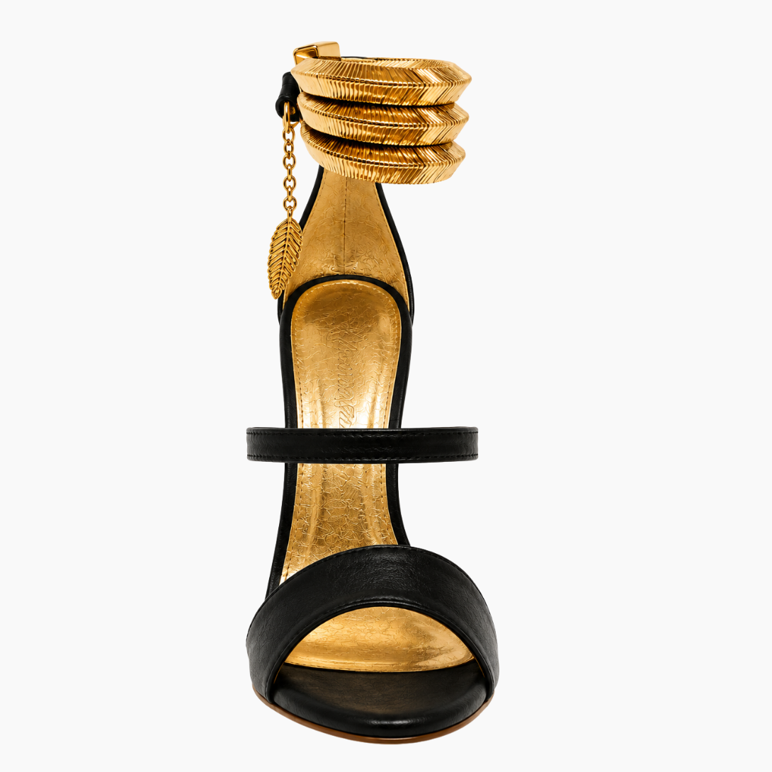 A single high-heeled sandal with a black leather open toe and ankle strap, featuring a textured gold insole, gold heel, and a stacked gold anklet-style detail on the ankle with a small gold leaf charm, shown against a plain white background.