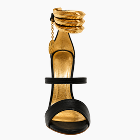 A single high-heeled sandal with a black leather open toe and ankle strap, featuring a textured gold insole, gold heel, and a stacked gold anklet-style detail on the ankle with a small gold leaf charm, shown against a plain white background.