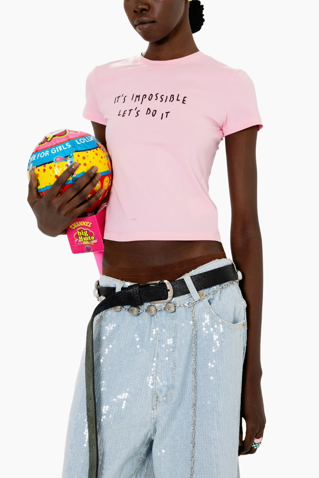 Person wearing a pink t-shirt with text, holding a colorful ball, and wearing sequin pants.