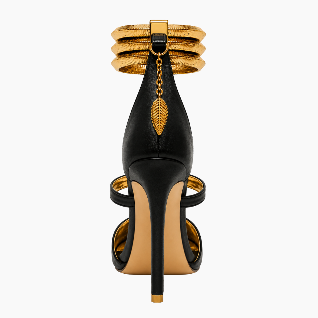 A single black leather high-heeled sandal with gold metallic accents, shown from the back on a light gray background. The shoe features a stiletto heel, a gold chain with a leaf charm, and stacked gold ankle cuffs.