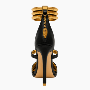 A single black leather high-heeled sandal with gold metallic accents, shown from the back on a light gray background. The shoe features a stiletto heel, a gold chain with a leaf charm, and stacked gold ankle cuffs.