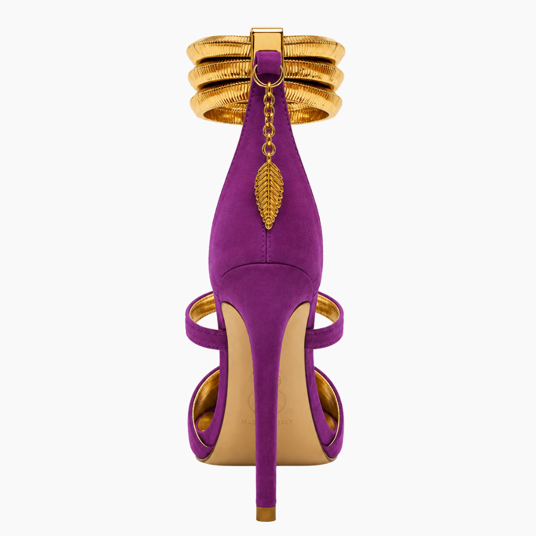 A single purple suede high-heeled sandal with gold metallic accents, including a multi-band ankle strap, a hanging leaf charm, and a gold-toned stiletto heel, shown against a plain white background.