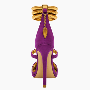 A single purple suede high-heeled sandal with gold metallic accents, including a multi-band ankle strap, a hanging leaf charm, and a gold-toned stiletto heel, shown against a plain white background.