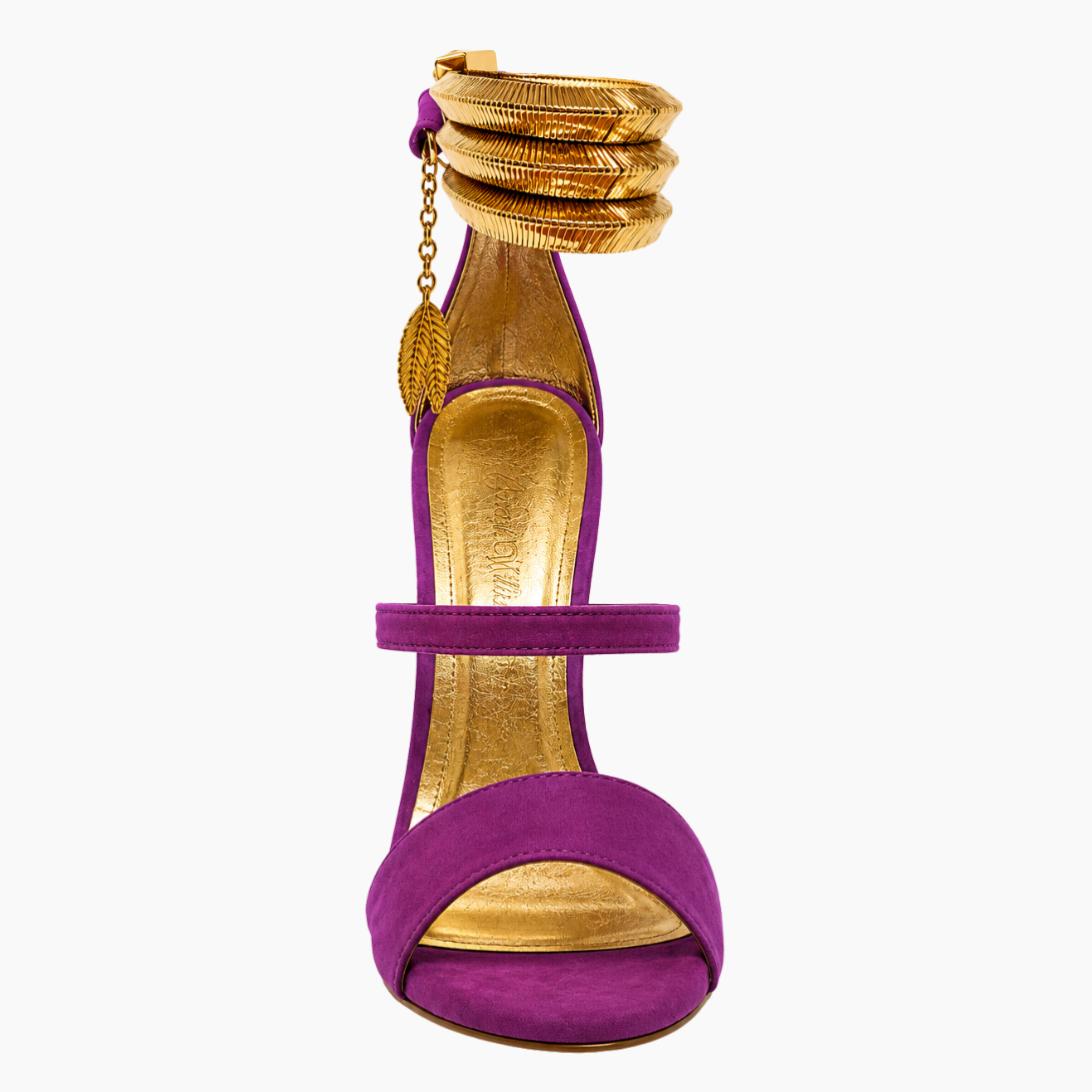 A purple suede high-heeled sandal with gold metallic accents, featuring a stacked gold ankle strap with a leaf charm and a gold insole, shown against a plain white background.