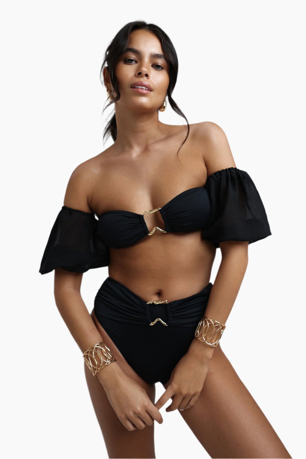Woman wearing a black off-shoulder top and high-waisted shorts with gold accessories on a white background