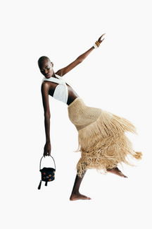 Model wearing a stylish outfit with a white top and beige skirt, holding a black handbag 