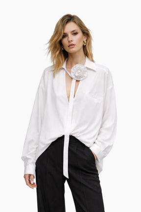 Woman wearing a white blouse with a floral accessory and black pants on a white background