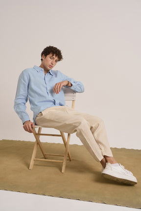 Man sitting on a chair wearing a light blue shirt, beige pants, and white sneakers.