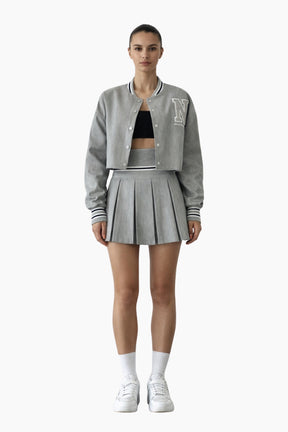 Person wearing a gray varsity jacket, black crop top, and pleated skirt on a white background