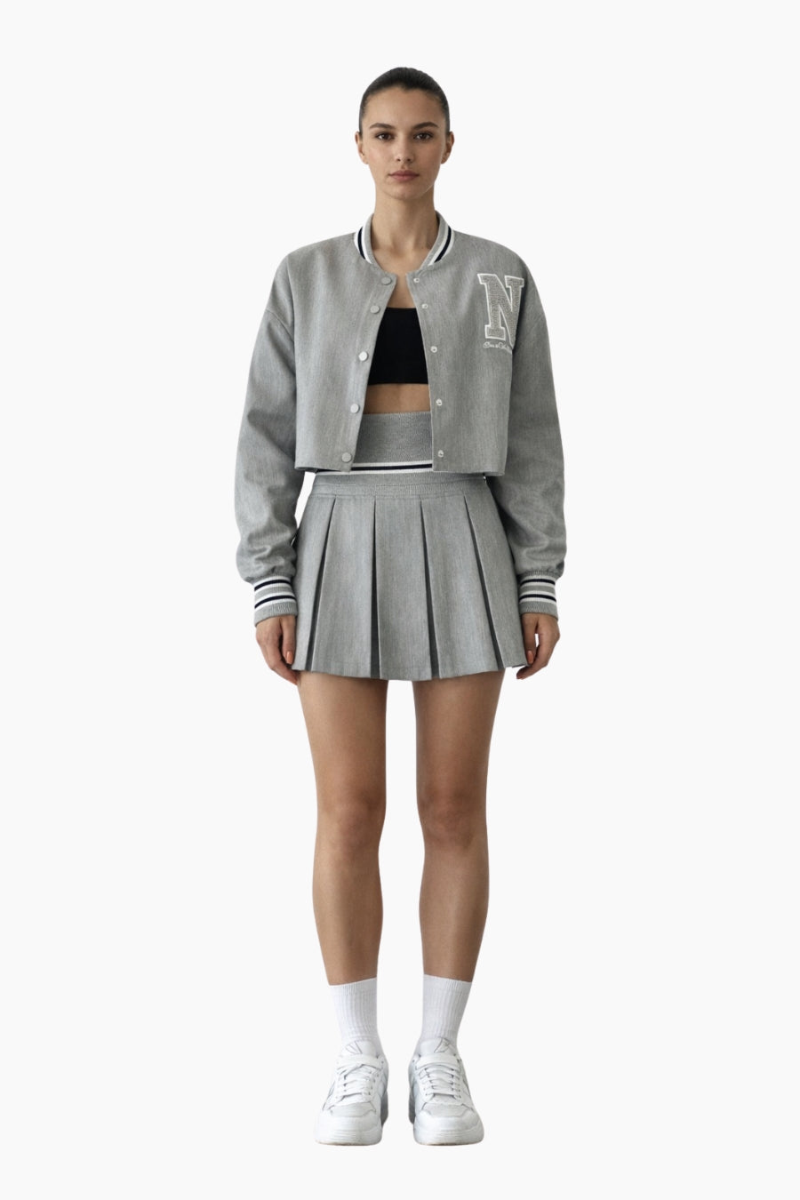 Person wearing a gray varsity jacket, black crop top, and pleated skirt on a white background