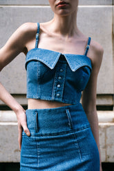 A woman wearing a blue denim shirt bustier with two collar details and shirt placket, paired with sunglasses and a denim skirt.