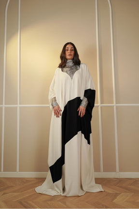 Woman wearing a white and black dress with a patterned shawl against a neutral wall.