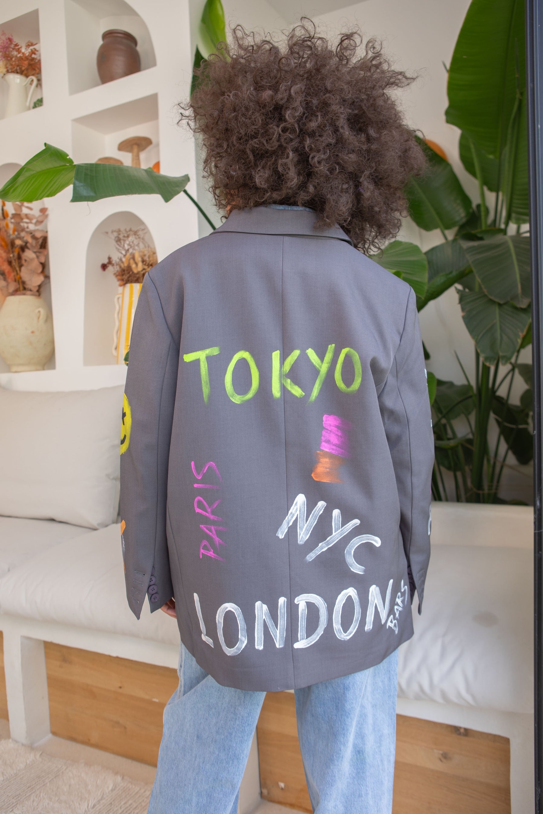Person wearing a gray jacket with 'TOKYO', 'PARIS', 'NYC', and 'LONDON' written on it, standing in a room with plants and shelves.