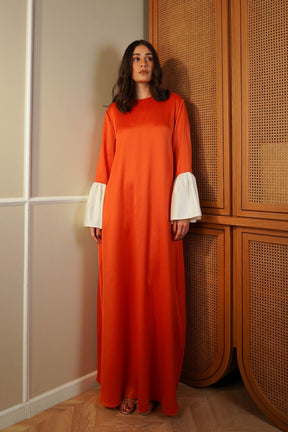 Woman wearing an orange dress with white cuffs standing against a wooden paneled wall.