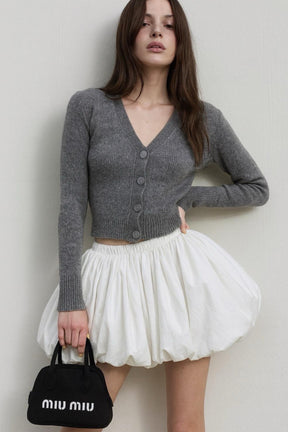 Woman wearing a gray cardigan and white skirt holding a black handbag with 'Miu Miu' branding against a plain background.