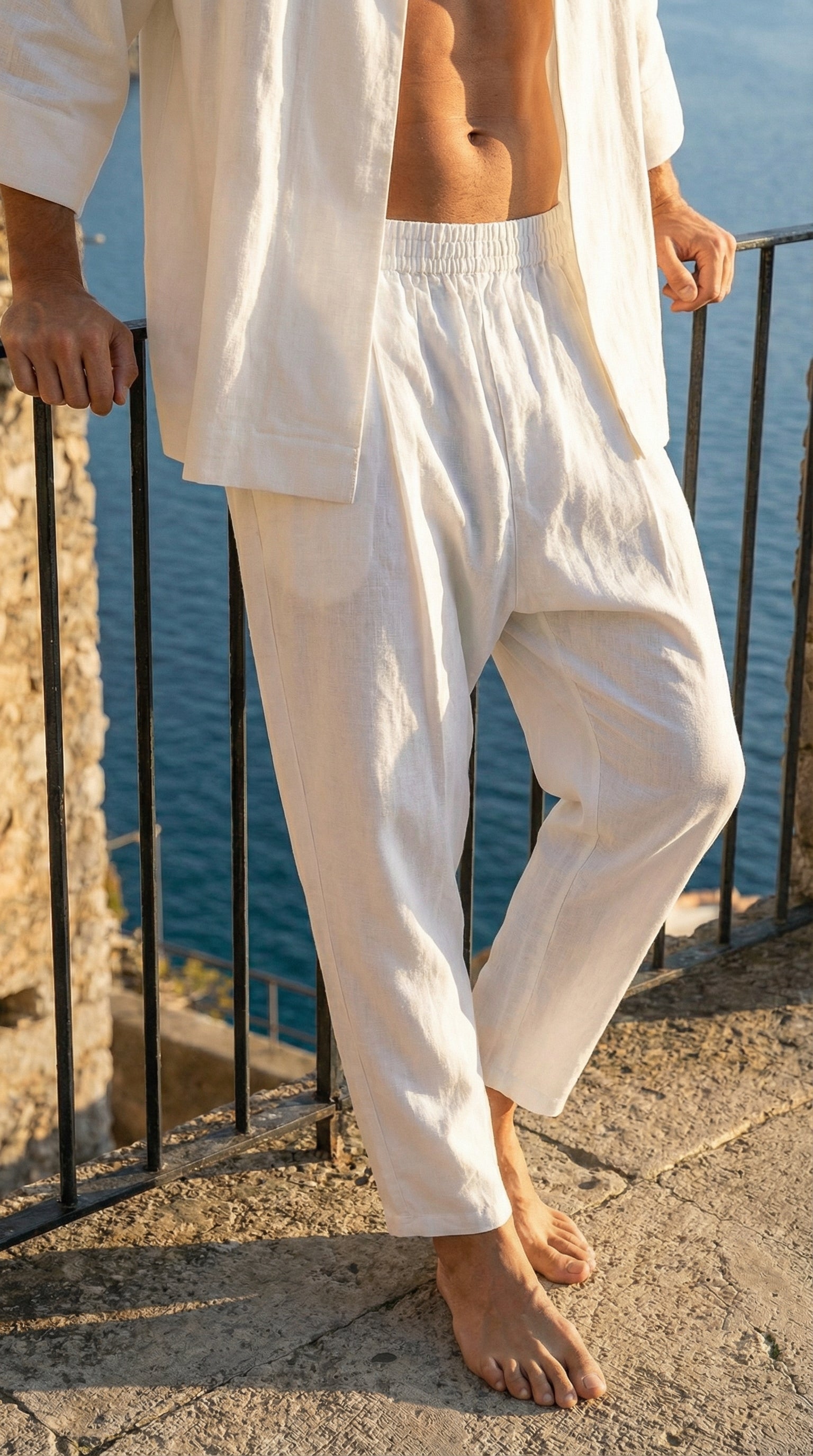Person wearing a loose-fitting white outfit standing on a stone ledge with water in the background.
