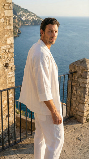 Man in white outfit standing on a stone balcony overlooking the ocean.