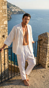 Man in white linen suit standing on a stone ledge overlooking the ocean.