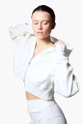 a white cropped hoodie featuring a structured silhouette with dropped shoulders, ribbed texture, and a statement front zipper pull paired with a short white skirt.