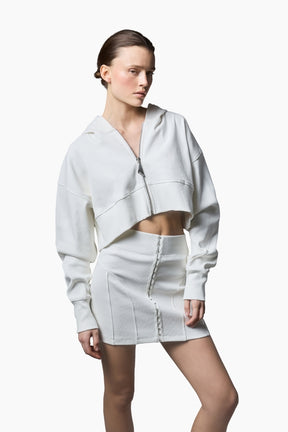 a white cropped hoodie featuring a structured silhouette with dropped shoulders, ribbed texture, and a statement front zipper pull paired with a short white skirt.