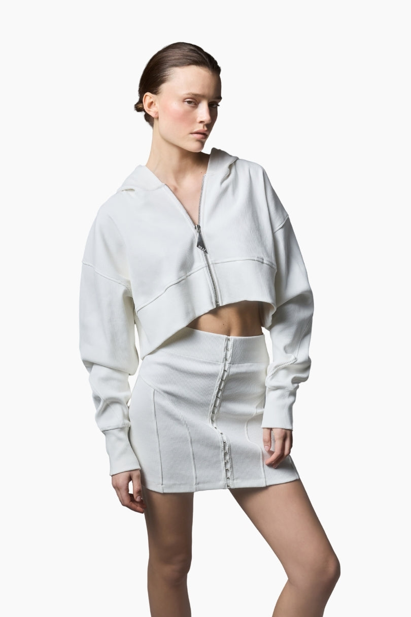 a white cropped hoodie featuring a structured silhouette with dropped shoulders, ribbed texture, and a statement front zipper pull paired with a short white skirt.