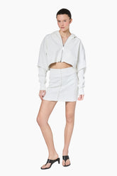 a white cropped hoodie featuring a structured silhouette with dropped shoulders, ribbed texture, and a statement front zipper pull paired with a short white skirt.