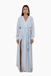 Woman wearing a light blue patterned dress with a slit. Flared floor-length dress with a V-neckline, balloon sleeves, two side slits, and a waist belt.