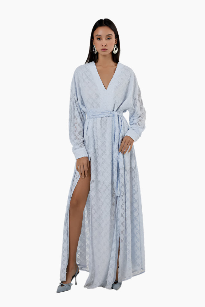 Woman wearing a light blue patterned dress with a slit. Flared floor-length dress with a V-neckline, balloon sleeves, two side slits, and a waist belt.