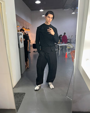 A person standing in a mirror wearing a black wrap shirt and black pants, with a minimalist interior visible in the background.