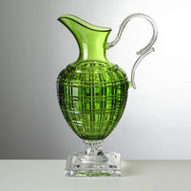A green acrylic pitcher with a geometric pattern and a silver handle and base, placed on a grey surface.