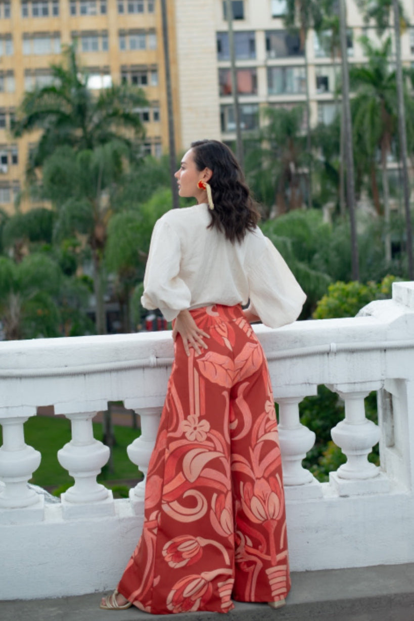 Woman in a white top and red patterned pants standing on a balcony with greenery and buildings in the background.