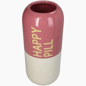 A capsule-shaped ceramic vase with a glossy dusty rose pink upper half and an off-white lower half, featuring the words 'HAPPY PILL' printed vertically in gold lettering on the pink section.
