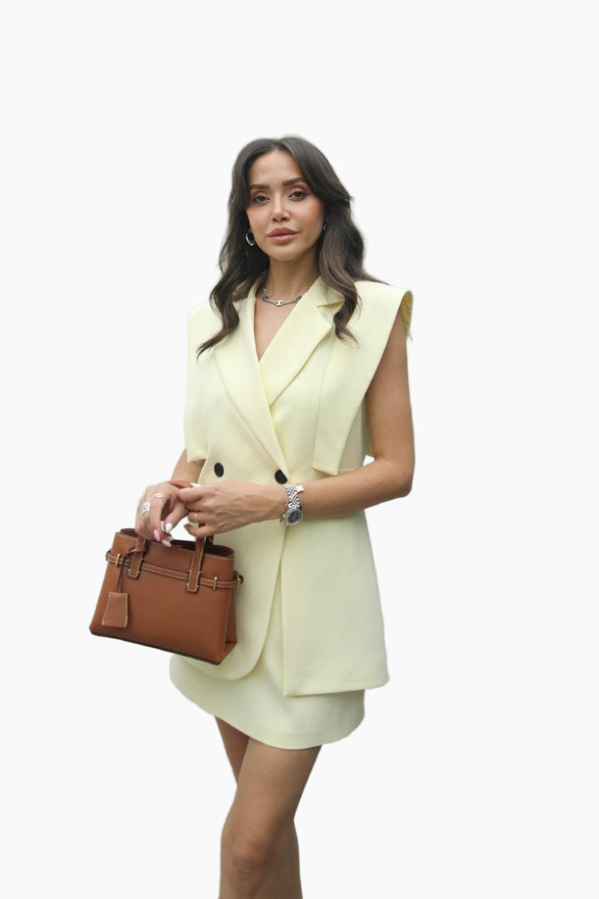 Woman wearing a light yellow sleeveless blazer dress holding a brown handbag on a white background
