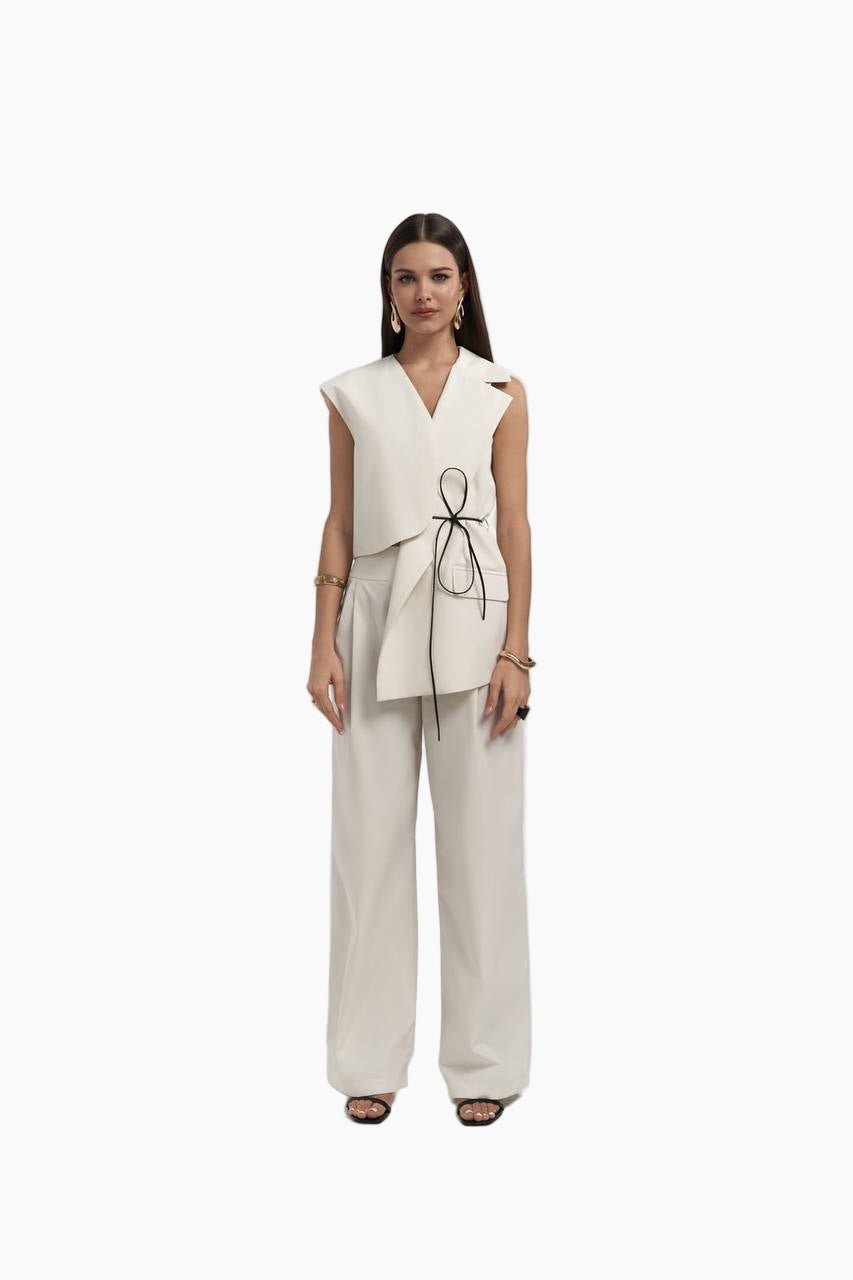 Woman wearing a white sleeveless top with a bow detail and beige pants on a white background