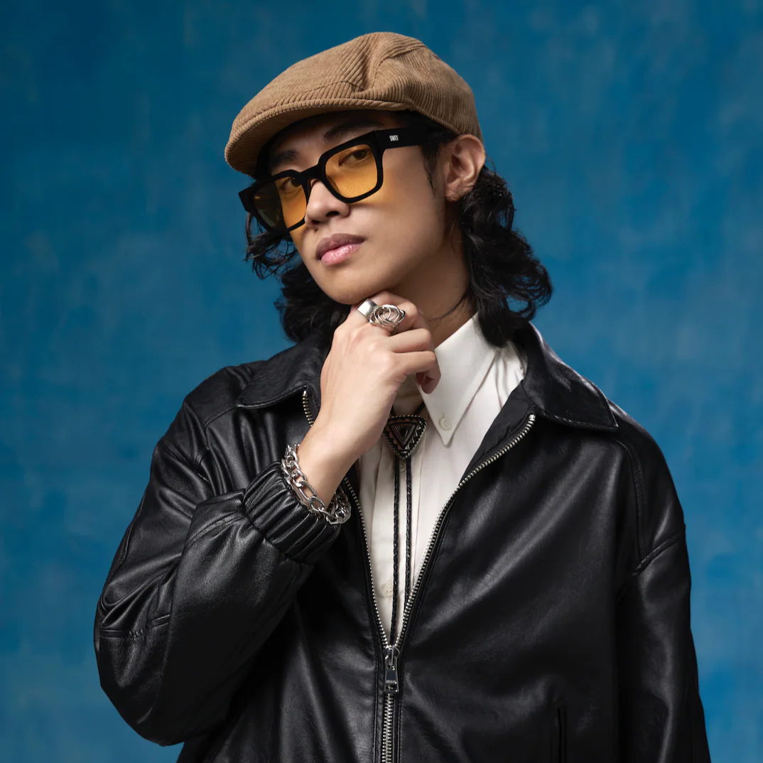 Person wearing a black leather jacket, beige cap, and yellow glasses against a blue background