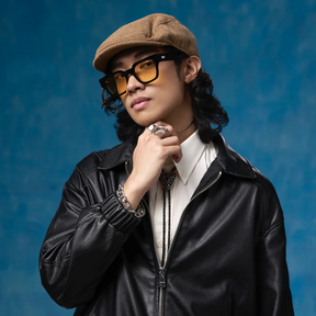Person wearing a black leather jacket, beige cap, and yellow glasses against a blue background