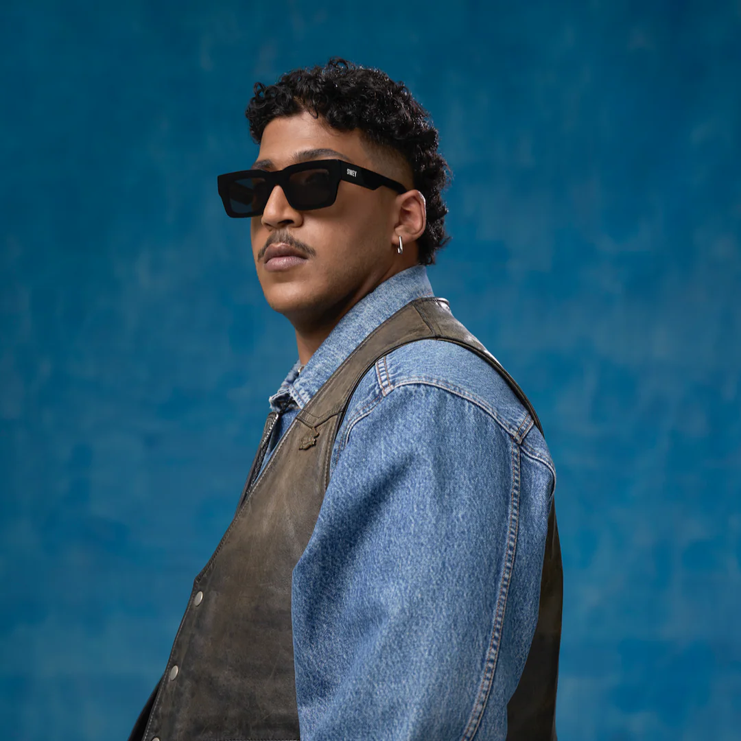 Man wearing a black sunglasses, a denim jacket, and leather vest against a blue background