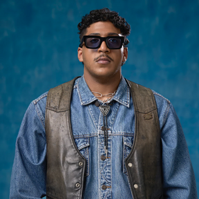 Man wearing sunglasses and a denim jacket with a leather vest against a blue background