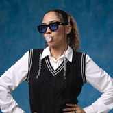 Person wearing sunglasses and a black sweater over a white shirt against a blue background