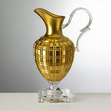 A ggold acrylic pitcher with a geometric pattern and a silver handle and base, placed on a grey surface.
